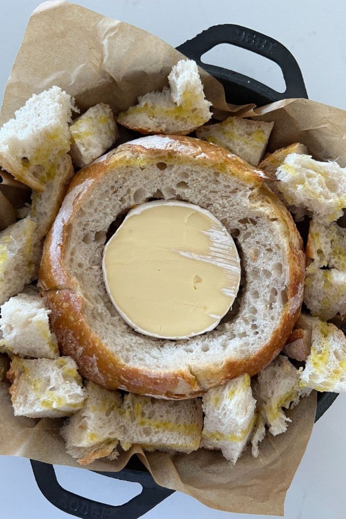 A sourdough bread bowl that has had the top cut out and a wheel of Brie cheese placed inside. There are chunks of sourdough bread sitting around the outside of the bread.