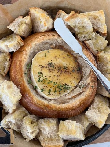 Sourdough bread bowl with gooey baked brie inside. The brie has been drizzled with honey and black pepper and fresh thyme leaves. There are chunks of toasted sourdough bread around the edges of the bowl and a silver knife in the photo.