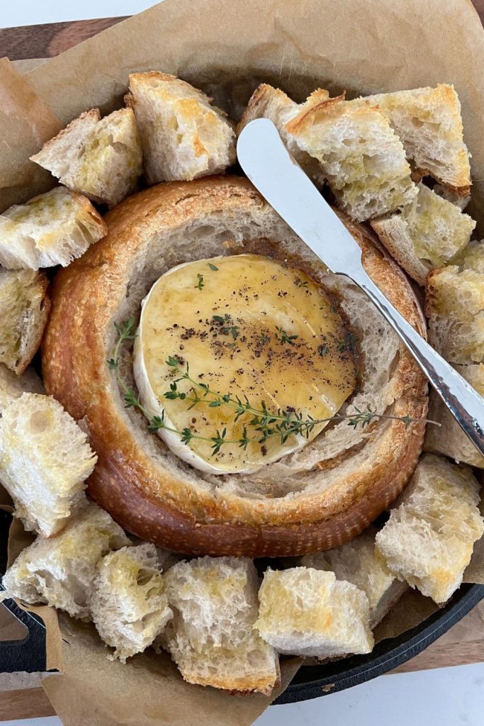 Sourdough bread bowl with gooey baked brie inside. The brie has been drizzled with honey and black pepper and fresh thyme leaves. There are chunks of toasted sourdough bread around the edges of the bowl and a silver knife in the photo.