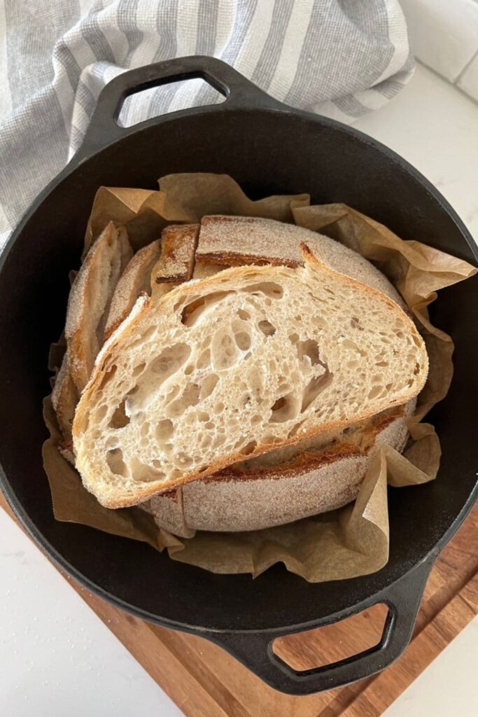 Slice of Dutch Oven Sourdough Bread sitting on a loaf that has been baked in a Lodge Cast Iron Dutch Oven. The Empty Dutch Oven can be seen in the background of the photo.