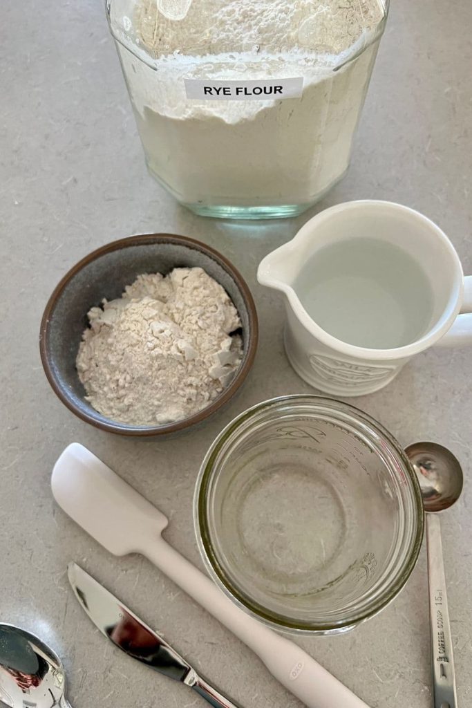 Photo shows a jar of flour, bowl of flour, small jug of water and a Ball Jar sitting on the counter top with a white jar spatula, silver spoon and knife.