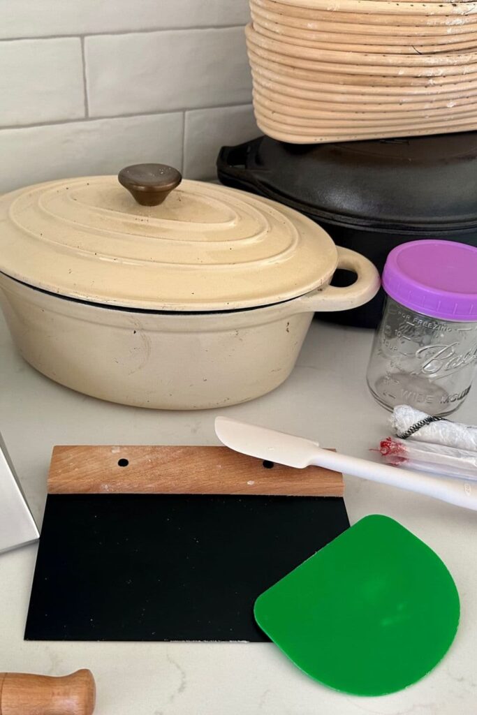 Black bench knife and green plastic dough scraper sitting with a white jar scraper. There is other sourdough baking equipment in the photo too including a cream cast iron Dutch Oven.