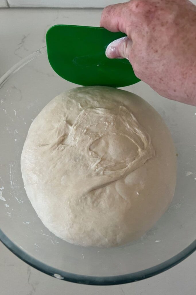 Sourdough bread dough being eased out of a glass bowl with a green plastic dough scraper.