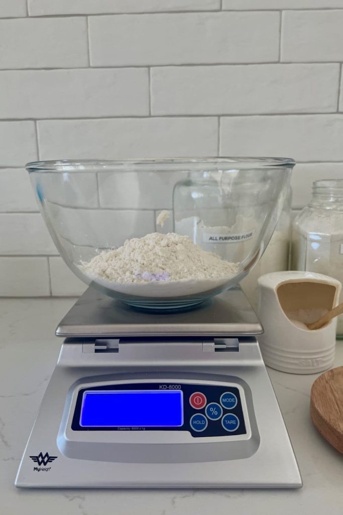 KD-8000 Baking Scale shown in the photo with a glass bowl of flour sitting on it. There is a salt pig and flour jar next to the scale and a white tiled wall in the background.