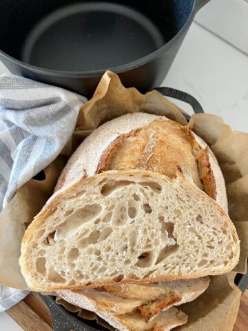 Slice of Dutch Oven Sourdough Bread sitting on a loaf that has been baked in a Lodge Cast Iron Dutch Oven. The Empty Dutch Oven can be seen in the background of the photo.