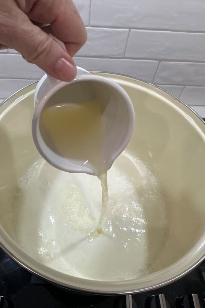 Pouring a jug of lemon juice into a pot of warm milk to make ricotta cheese at home.