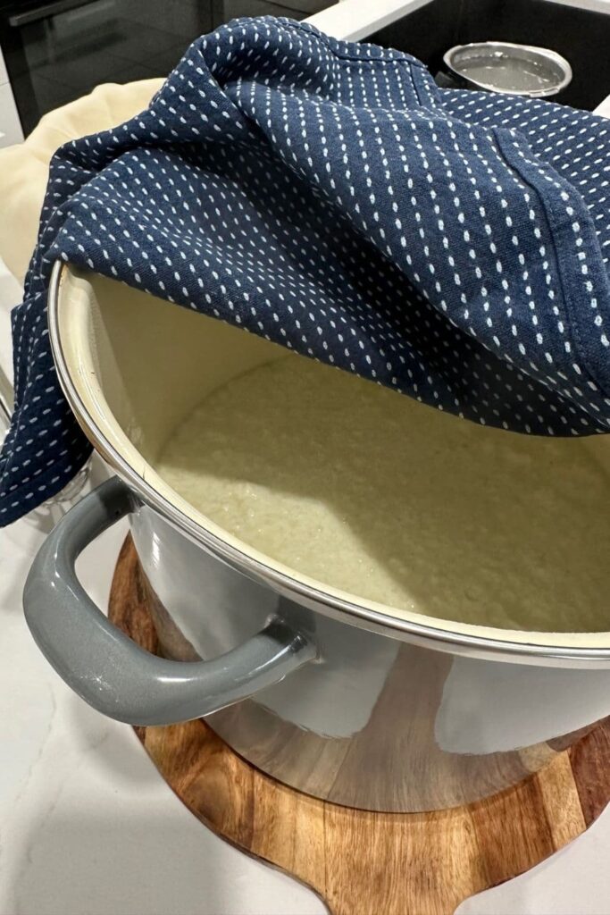 A pot of ricotta cheese sitting on a wooden board, covered with a navy blue dish cloth.