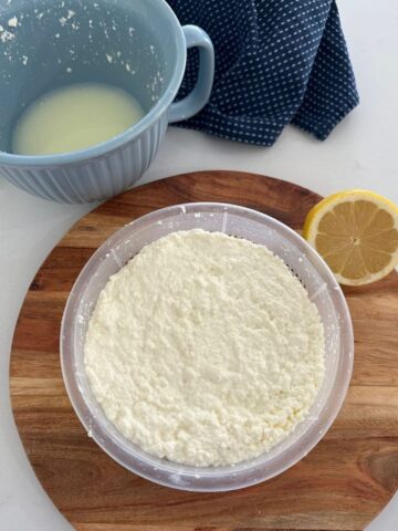 Ricotta cheese that has been strained in a plastic ricotta basket. The basket is sitting on a wooden board and there is a lemon to the right and a blue jug of whey on the left.