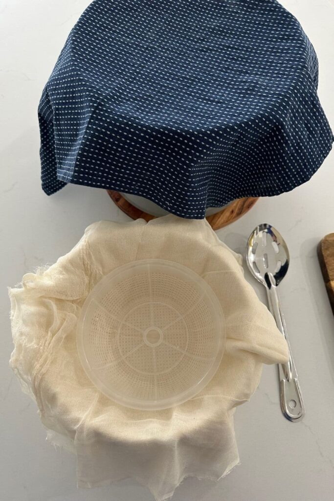 A blue cotton dish towel covering a pot of ricotta that is being left to sit. There is also a plastic ricotta basket, slotted spoon and cheesecloth in the photo.