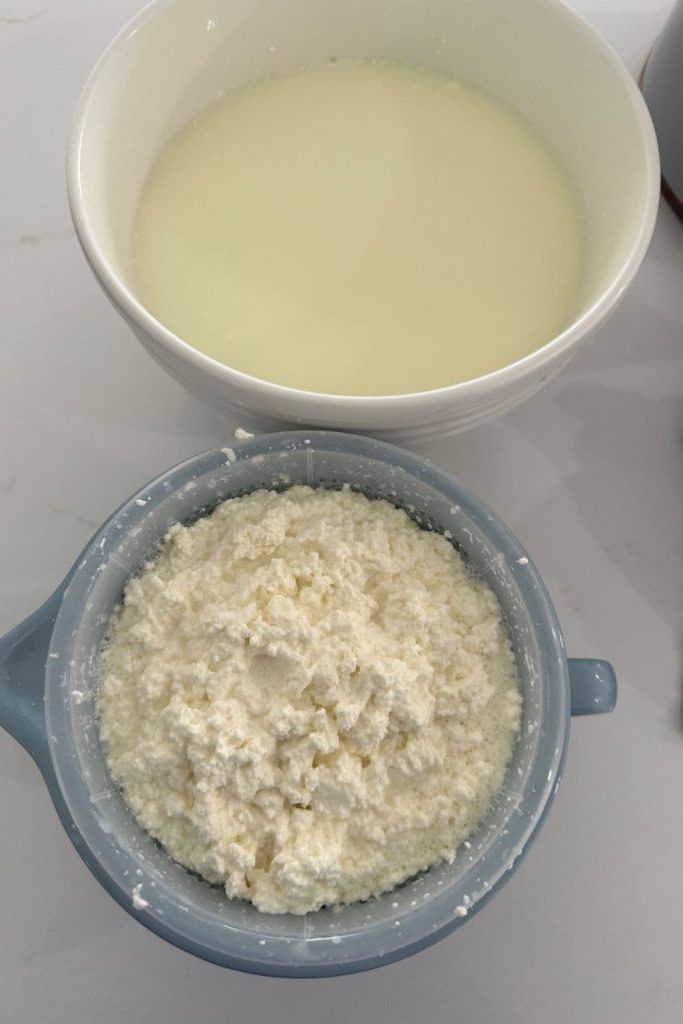 A big bowl of ricotta whey sitting behind a small blue jug with a plastic basket of homemade ricotta draining into it.
