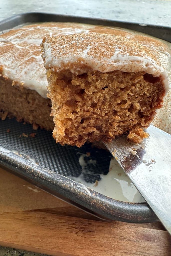 Sourdough applesauce cake with Greek Yogurt Glaze. There is a close up of a slice of cake being lifted out of the tin and you can see the rest of the cake in the background of the photo.
