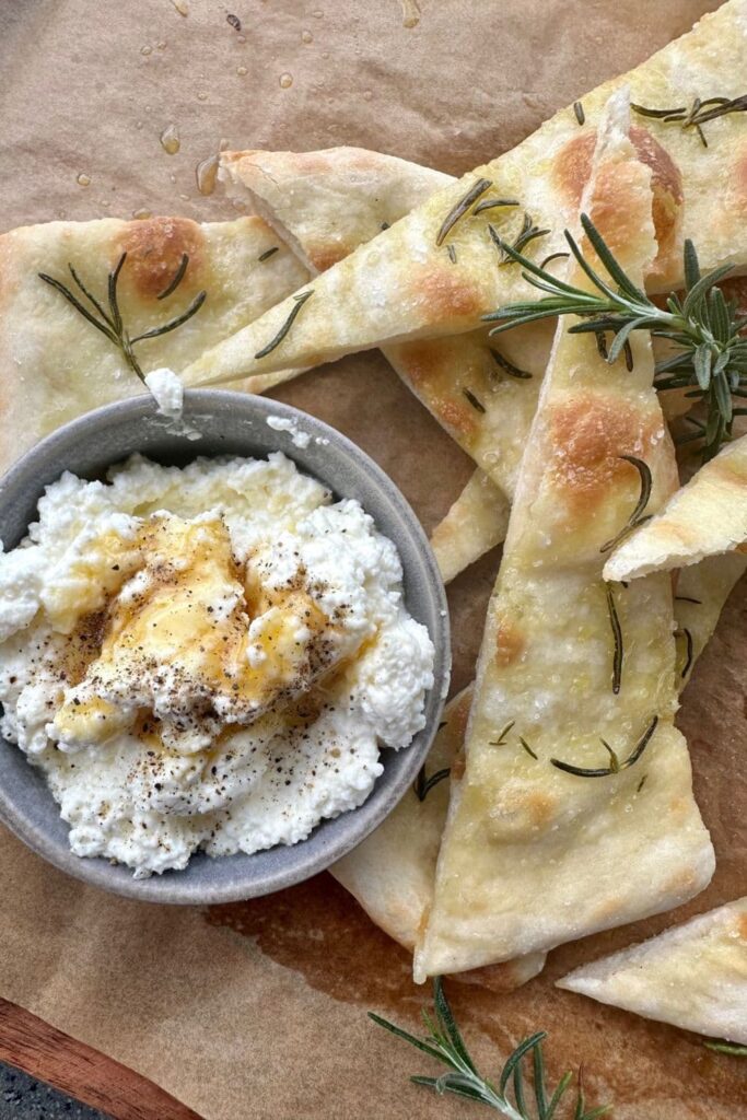 Bowl of fresh ricotta drizzled with honey and served next to sourdough pizza bianca on a wooden board.