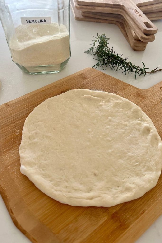 Sourdough pizza bianca dough shaped onto a wooden pizza peel. There is some fresh rosemary and a jar of semolina in the background.