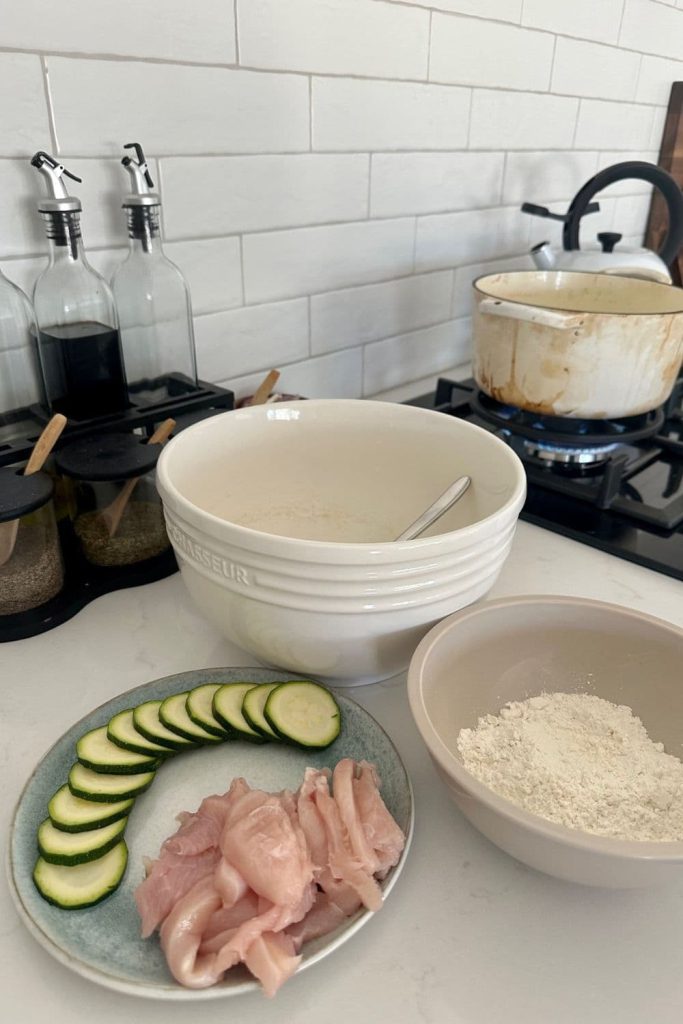 A plate of chicken and zucchini slices sitting next to a bowl of sourdough discard batter and bowl of flour. There is a pot of vegetable oil on the stove ready for deep frying.