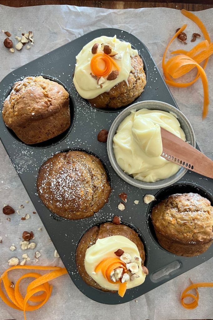 Sourdough carrot cake muffins sitting in a muffin tray. There are slivers of carrot around the tray and a little dish of cream cheese icing being spread on with a rose gold butter knife.