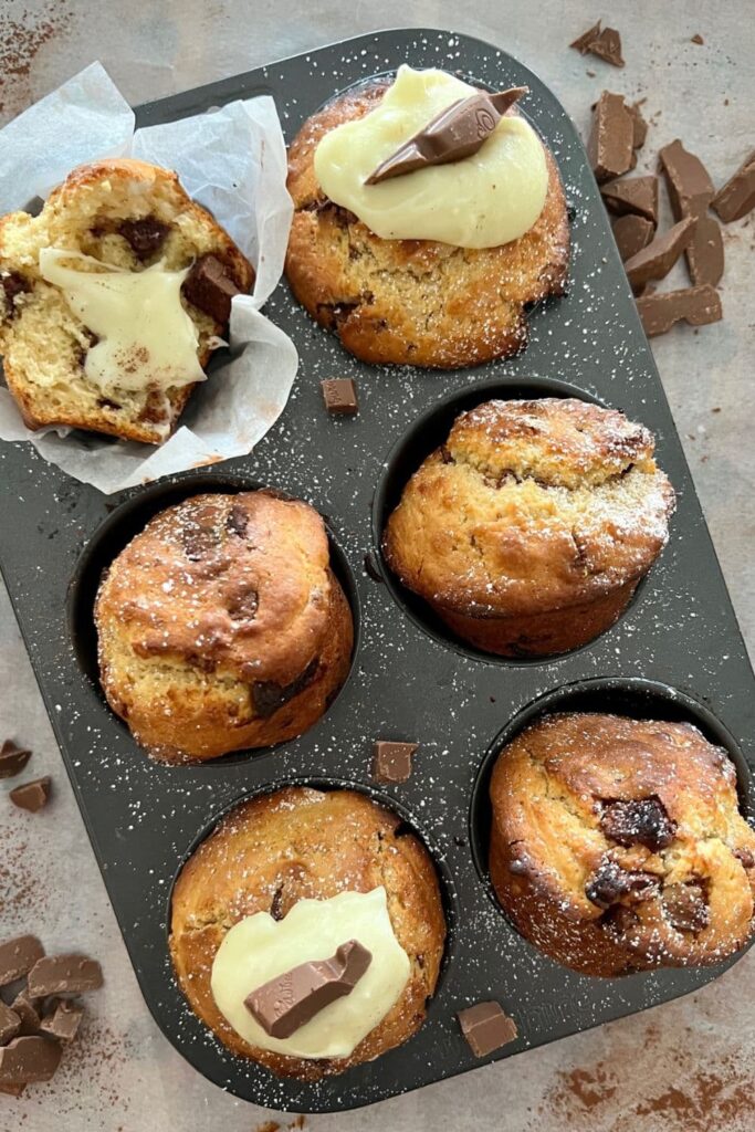 Sourdough chocolate chunk muffins sitting in a muffin tray. Some of the muffins have been topped with cream cheese.