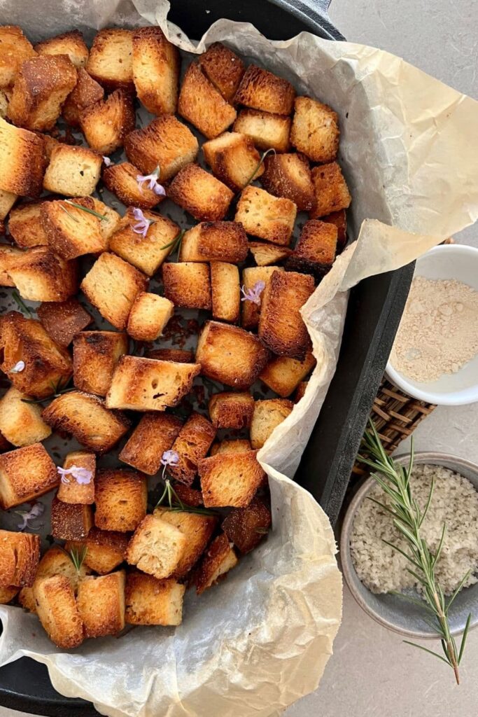 Crunchy sourdough croutons sitting in a cast iron tray. They have been topped with rosemary leaves and flowers.