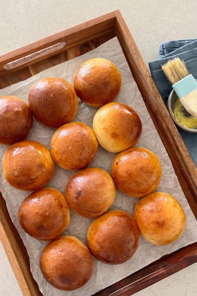 Tray of 12 sourdough brioche buns sitting next to a blue dish towel and pastry brush with butter.