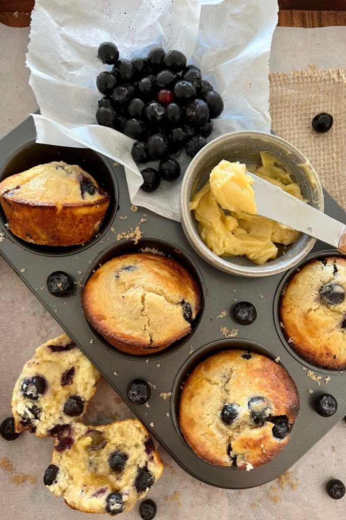 Overnight sourdough blueberry muffins displayed in a muffin tin. There is a bunch of blueberries in the background of the picture and one of the muffins is cut open so you can see inside the muffin.