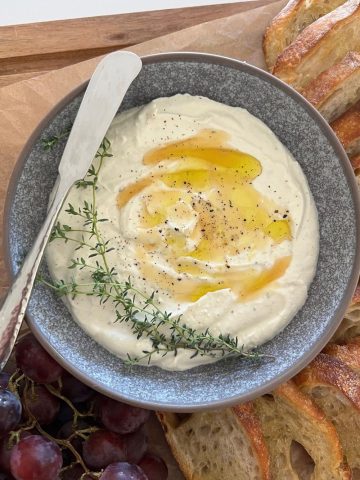 Whipped Ricotta Dip drizzled with honey and olive oil, dressed with fresh thyme leaves and served in a grey stoneware dish. There is a silver pate knife sitting on the edge of the bowl.