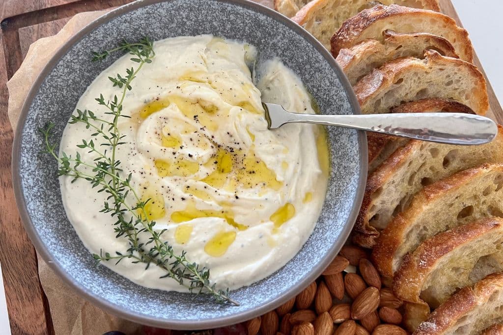 Whipped Ricotta Dip drizzled with honey and olive oil, dressed with fresh thyme leaves and served in a grey stoneware dish. There is a silver pate knife sitting on the edge of the bowl.