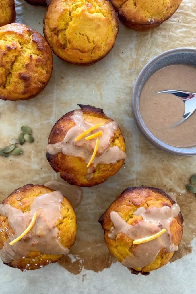 Sourdough pumpkin muffins sitting on a counter next to a bowl of orange maple glaze. Some of the glaze has been drizzled over the sourdough muffins.