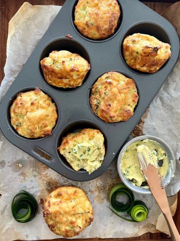 A tray of 6 sourdough zucchini muffins with cheese and chives sitting in a muffin tin. There is a bowl of whipped herb butter sitting next to the sourdough muffins.