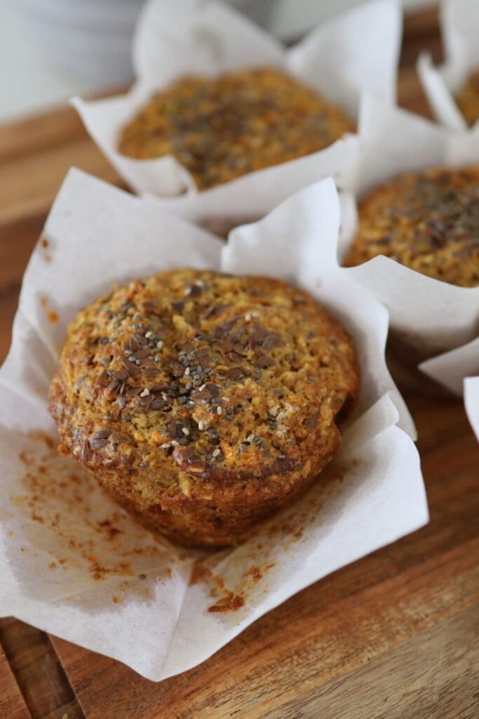 A close up of a sourdough morning glory muffin a white paper liner. You can see other muffins in the background of the photo.