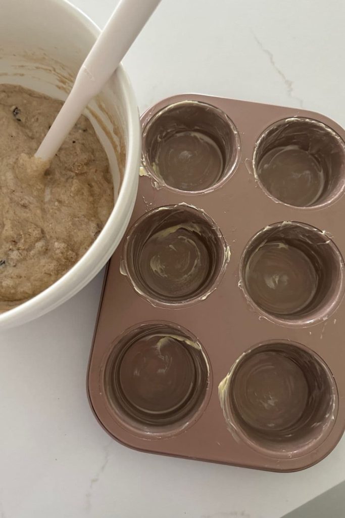 A rose gold six hole muffin tin that has been greased with butter. There is a bowl of muffin mixture sitting to the left of the muffin tin.