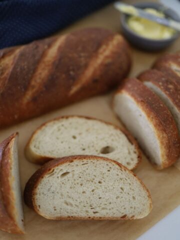 Sourdough French Bread that has been sliced and laid out on a piece of parchment paper. There is a dish of butter in the background of the photo.