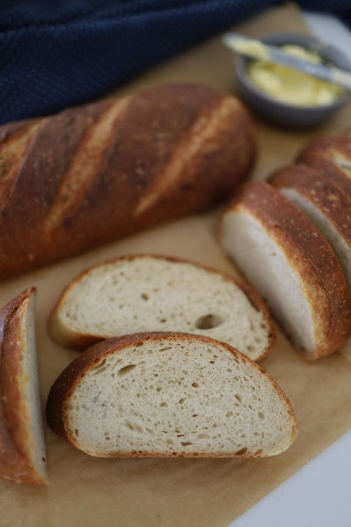 Sourdough French Bread that has been sliced and laid out on a piece of parchment paper. There is a dish of butter in the background of the photo.