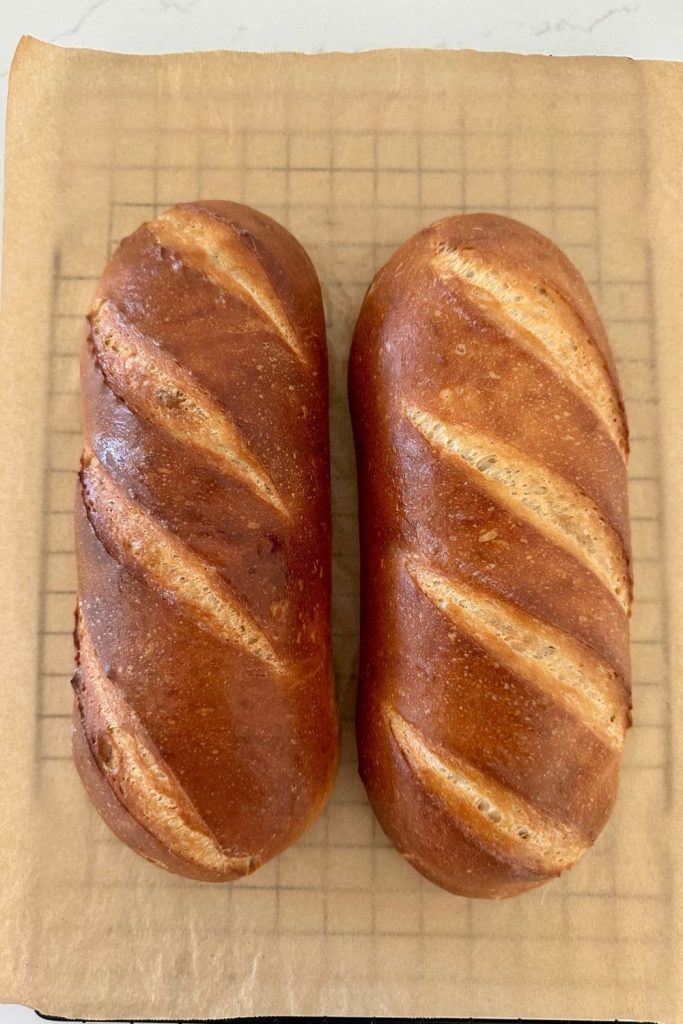 Two loaves of Sourdough French Bread fresh out of the oven. They are sitting on a piece of parchment paper on top of a black wire cooling rack.