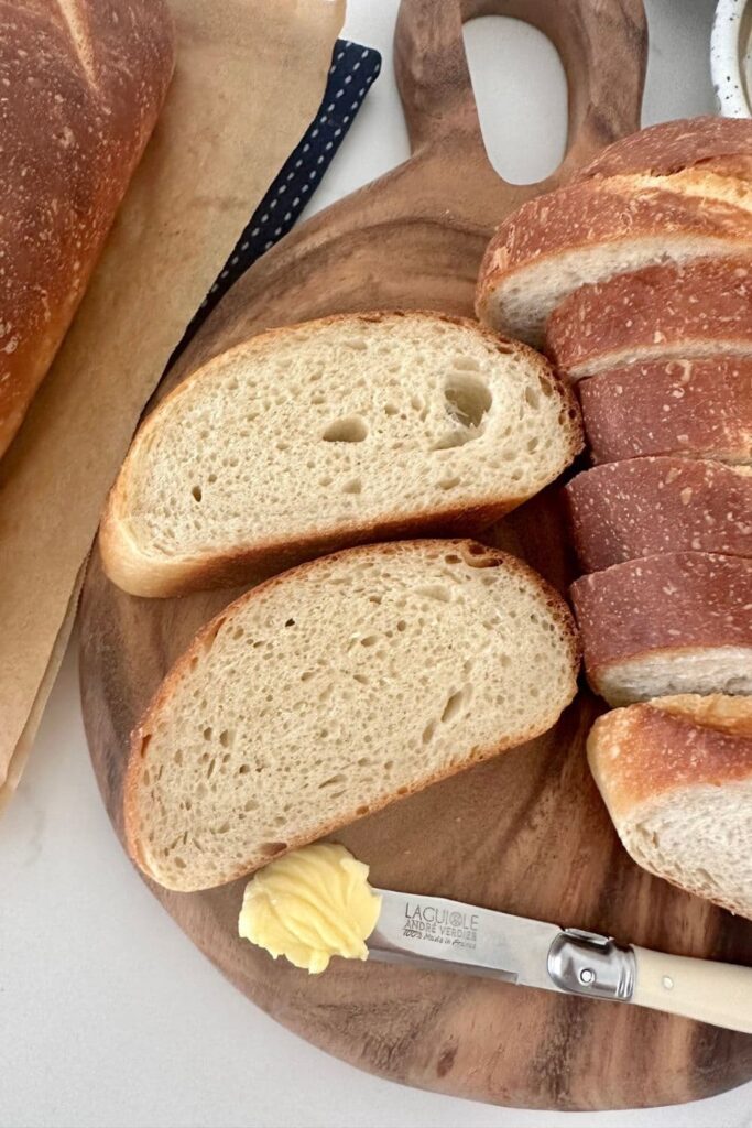 Slices of sourdough French Bread that are sitting on a wooden board with a small knife with butter on it.