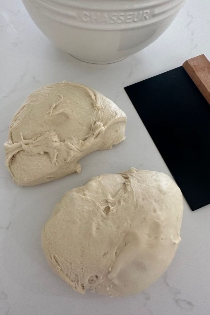 French bread dough that has been divided into two pieces. There is a black bench scraper and cream porcelain bowl in the photo.