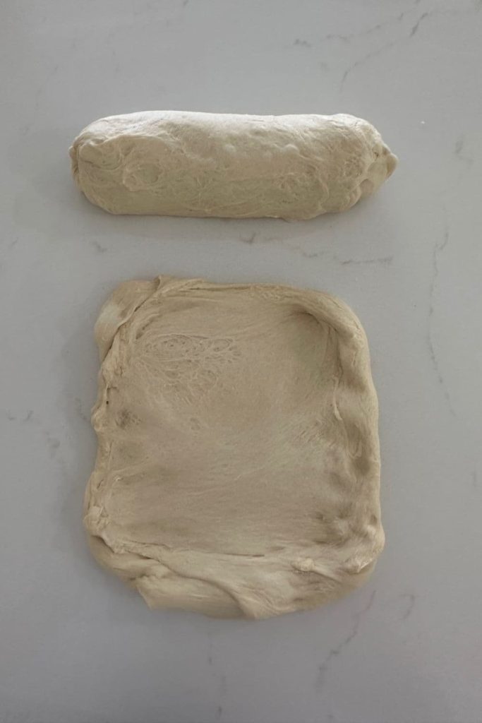 A piece of dough that has been shaped into a log sitting behind another piece of dough that has been pulled out into a rough rectangle to make sourdough French Bread.