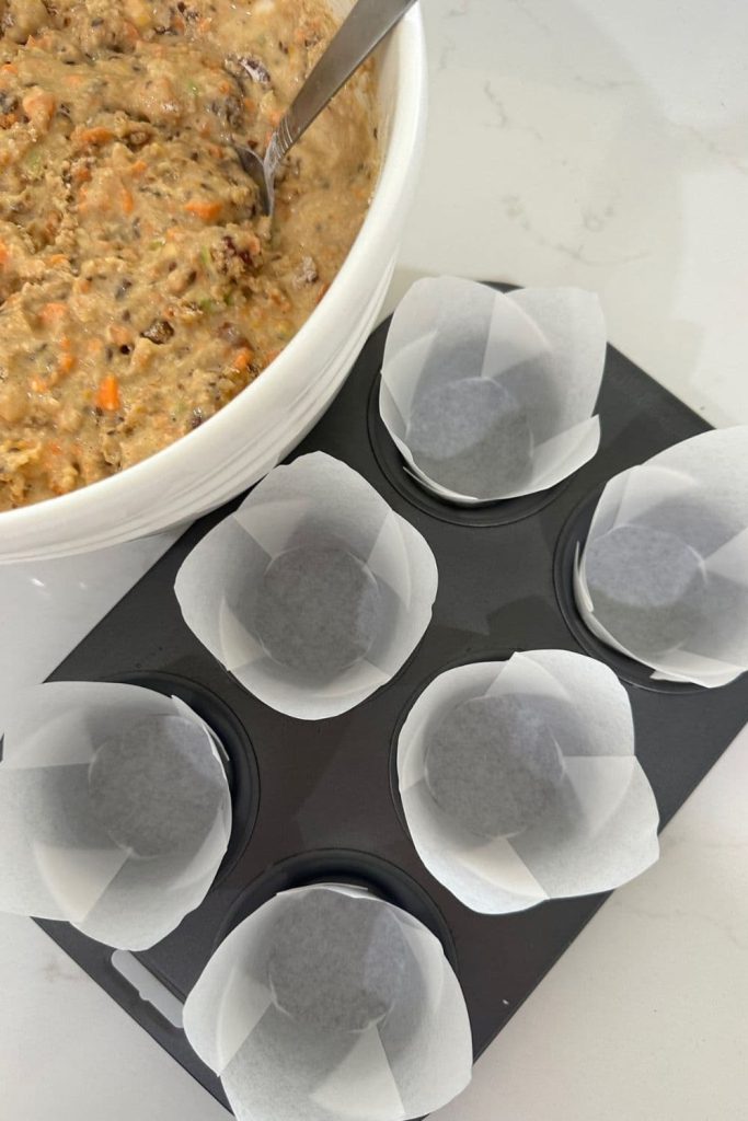 6 white paper liners in a 6 hole muffin tin sitting next to a bowl of sourdough muffin mixture.