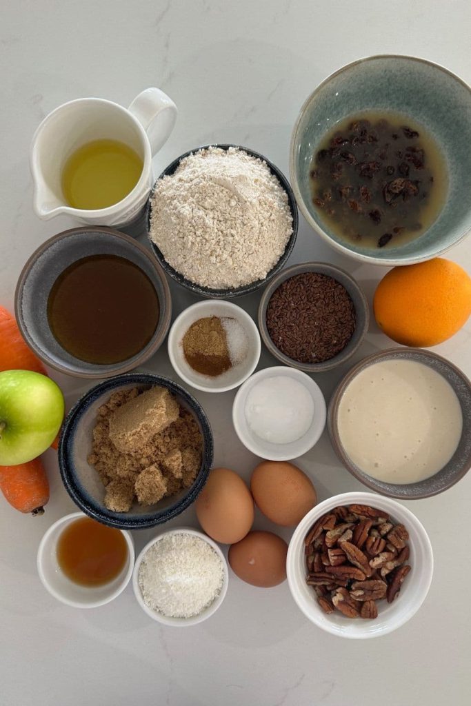 All of the ingredients used to make sourdough morning glory muffins including whole wheat flour, carrot, apple, pecans, eggs, seeds, orange, brown sugar.