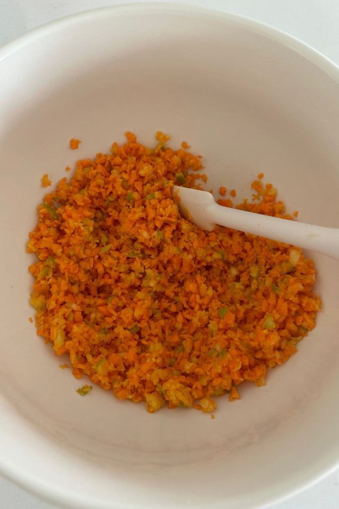 A bowl of grated carrot and apple being stirred together with a white jar spatula.