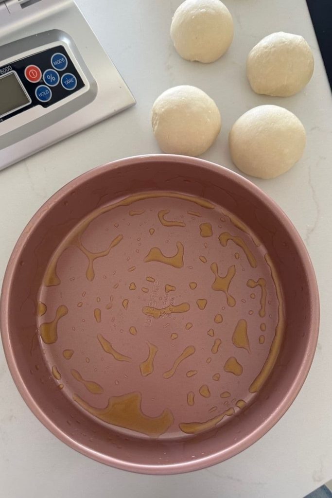 A round pink cake tin that has been coated in oil. There is a set of scales in the picture also and 4 dough balls above the cake tin.