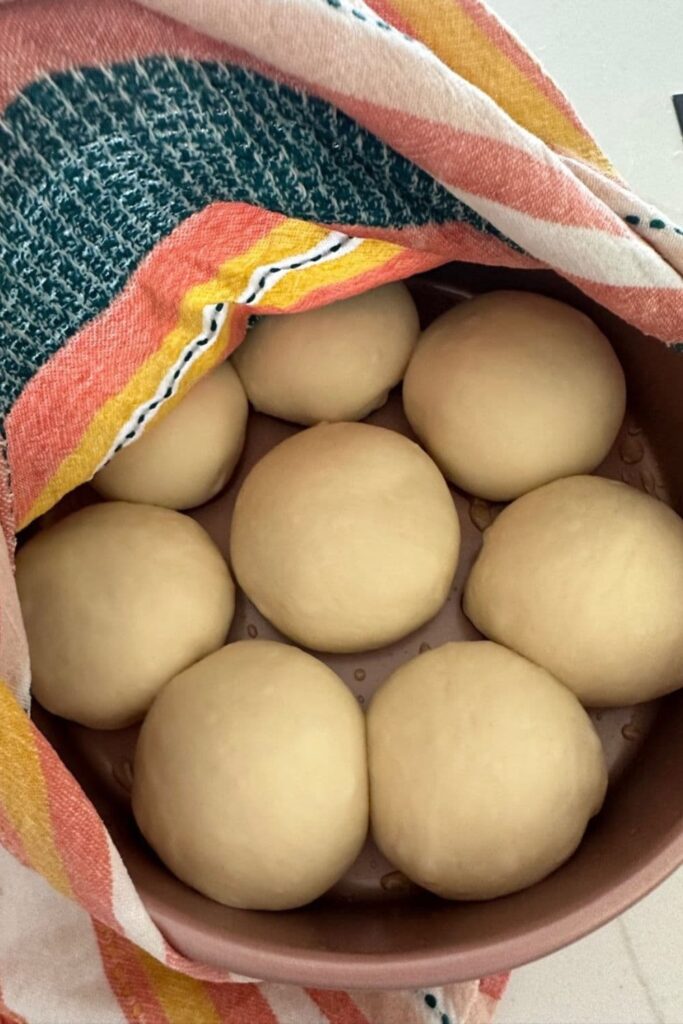 Dough balls sitting in a round pink cake tin that has been oiled. There is one in the middle and the other 7 are sitting around the outside of the tin. There is a colorful dish towel covering some of the dough.