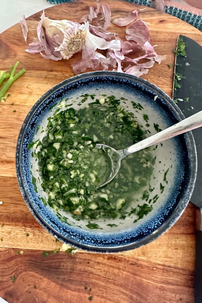 Herb and garlic oil in a blue edge stoneware bowl. You can see the garlic skin sitting behind the bowl and a knife used to chop the parsley to the right of the bowl.