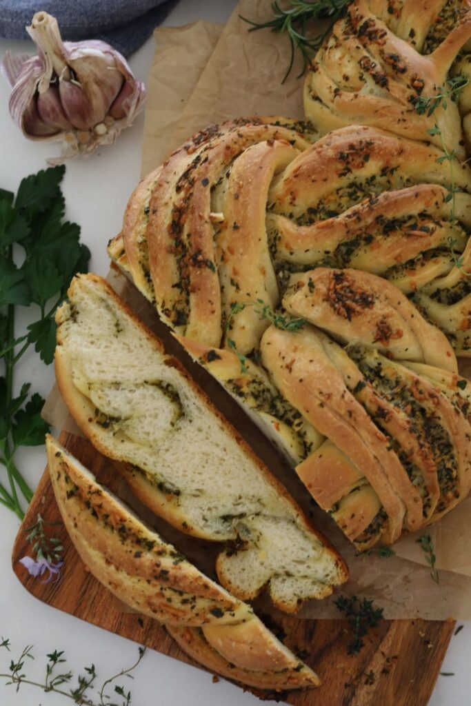 Sourdough Garlic & Herb Twist loaf sitting on a wooden board with some parchment paper. There is a demin blue dish towel in the background.