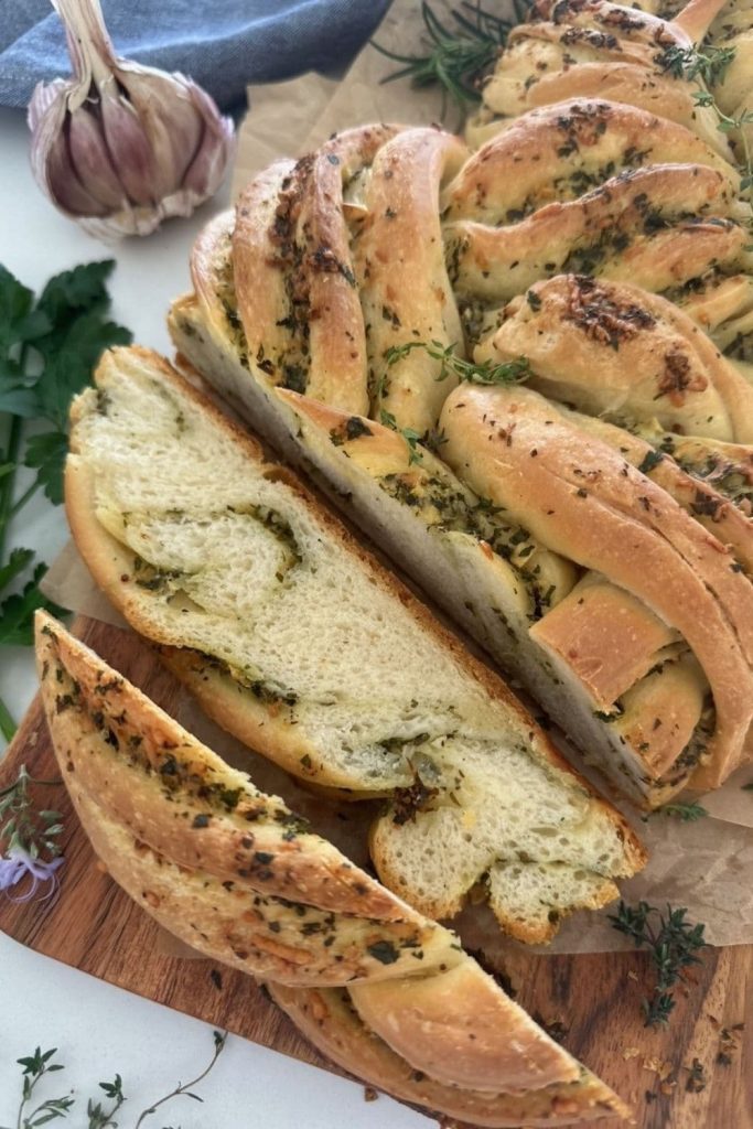 Sourdough Garlic & Herb Twist loaf sitting on a wooden board with some parchment paper. Two pieces have been sliced off the loaf and one is sitting so you can see the ribbons of herb and garlic butter swirled through the bread. There is a demin blue dish towel in the background.