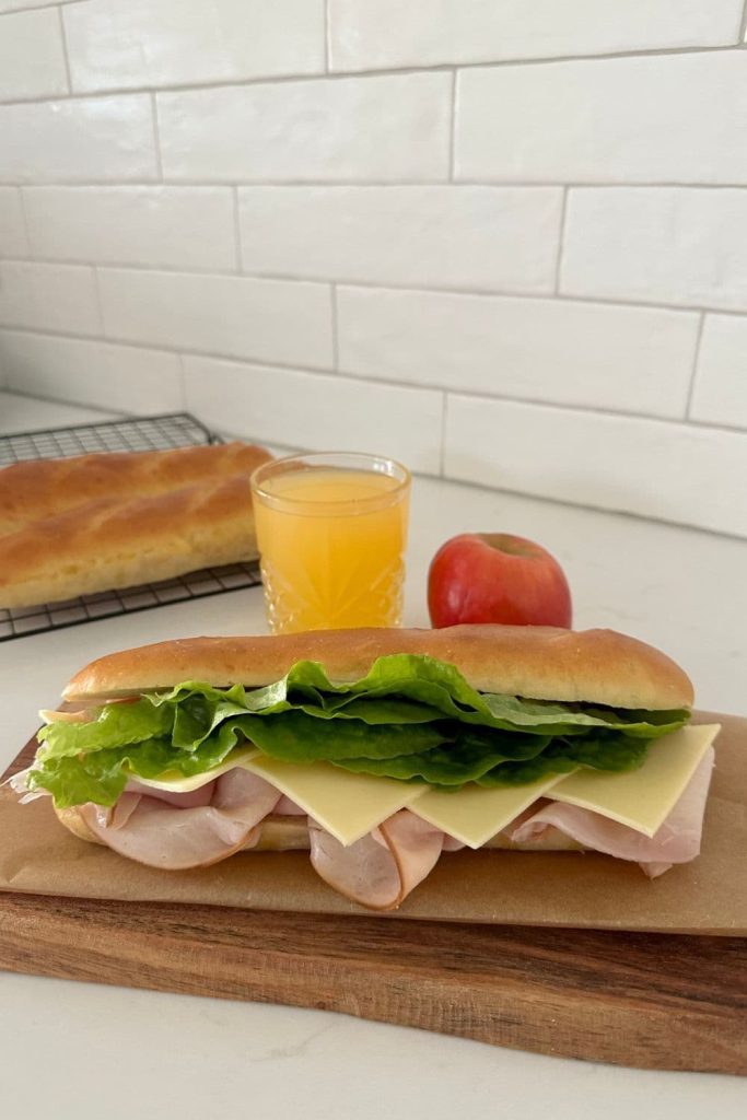 A sourdough sandwich roll filled with ham, cheese and lettuce. The sandwich is sitting in front of a glass of orange juice and an apple. There is a white tiled wall in the background.