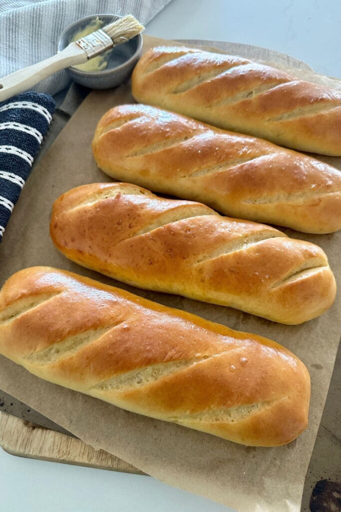 4 sourdough sandwich rolls sitting on a baking tray lined with baking paper. The sourdough subs have been brushed with butter.