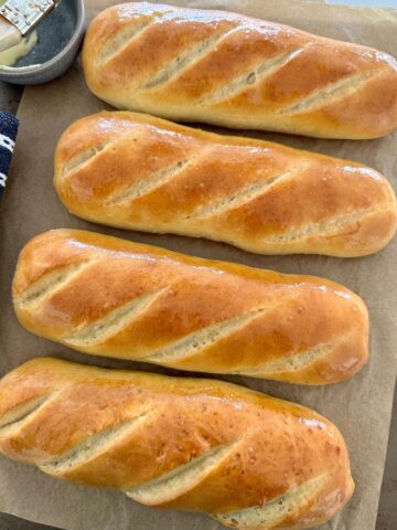 4 sourdough sandwich rolls sitting on a baking tray lined with baking paper. The sourdough subs have been brushed with butter.