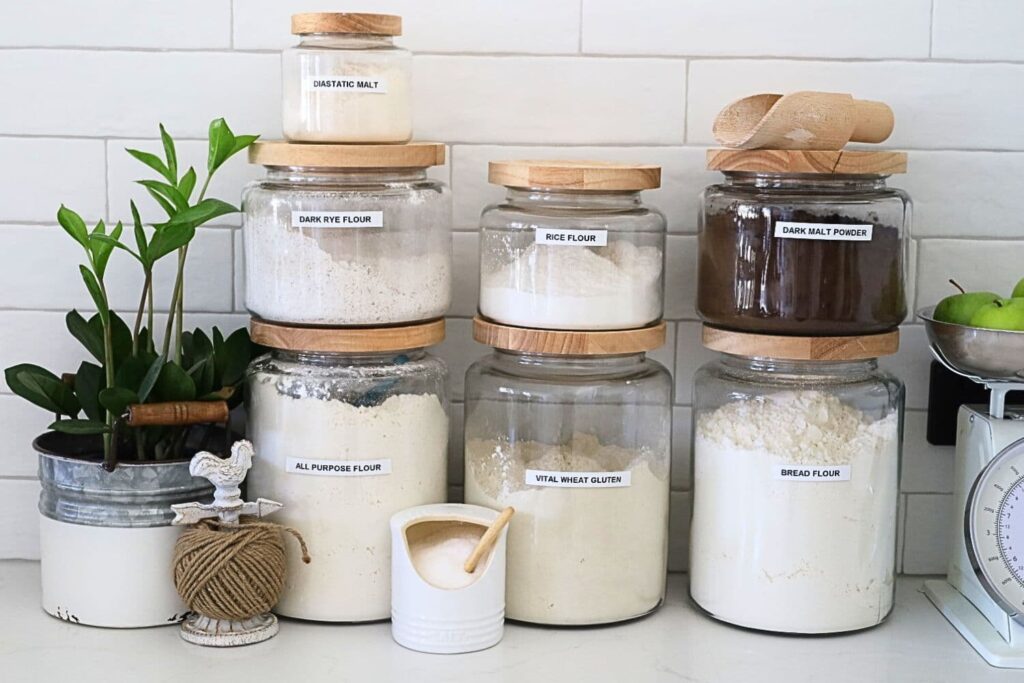 3 large jars of flour surrounded by some smaller jars containing rye flour, dark malt flour, diastatic malt and salt.