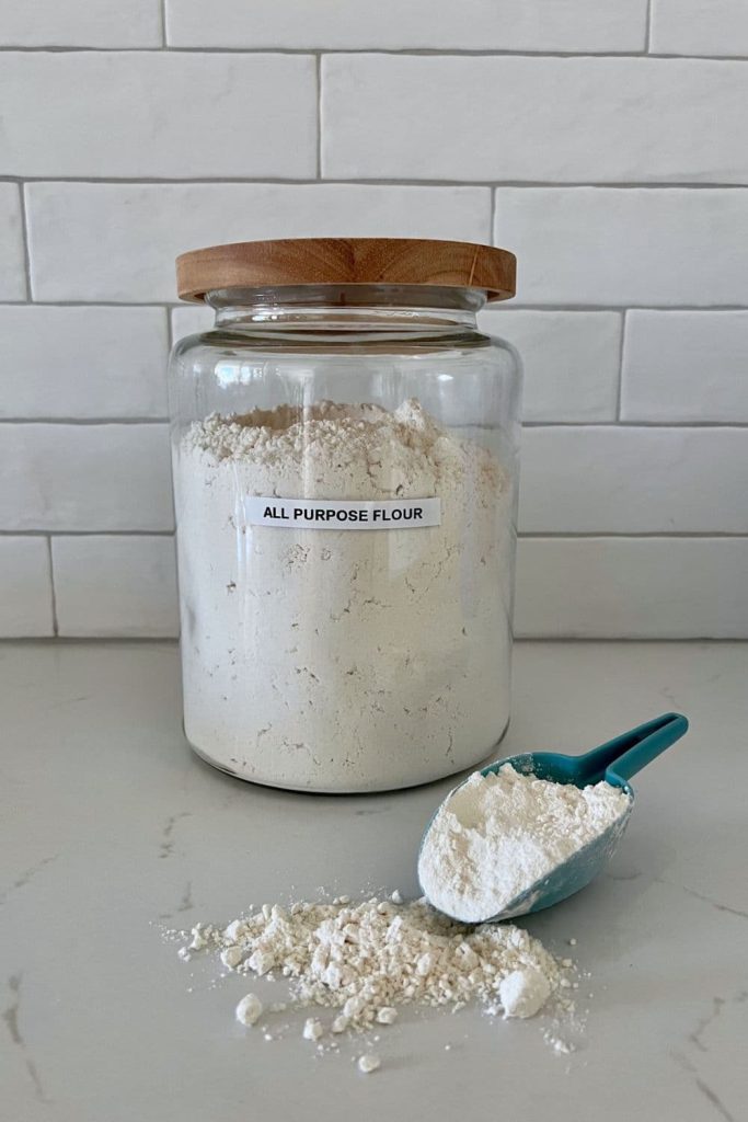 A large jar of all purpose flour sitting on a white kitchen counter next to a blue scoop.