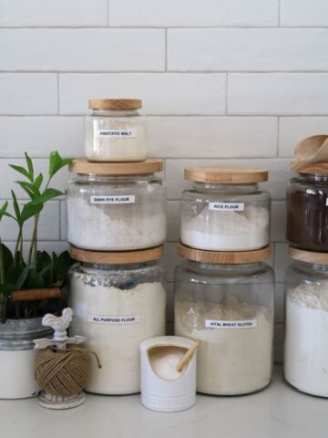 3 large jars of flour surrounded by some smaller jars containing rye flour, dark malt flour, diastatic malt and salt.