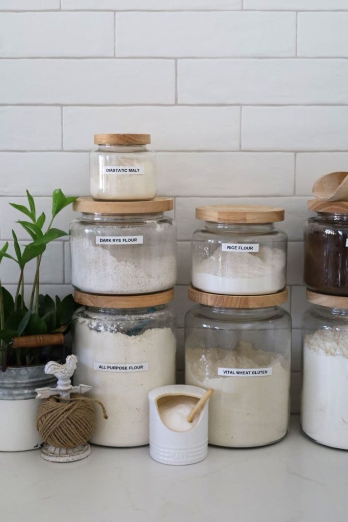 3 large jars of flour surrounded by some smaller jars containing rye flour, dark malt flour, diastatic malt and salt.